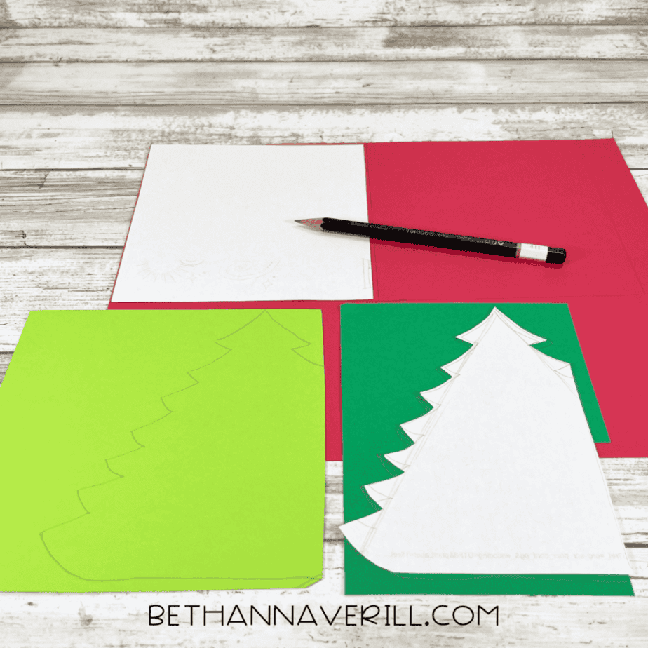 Christmas tree patterns traced onto green cardstock using a pencil, with white and red cardstock sheets nearby on a white wood surface.