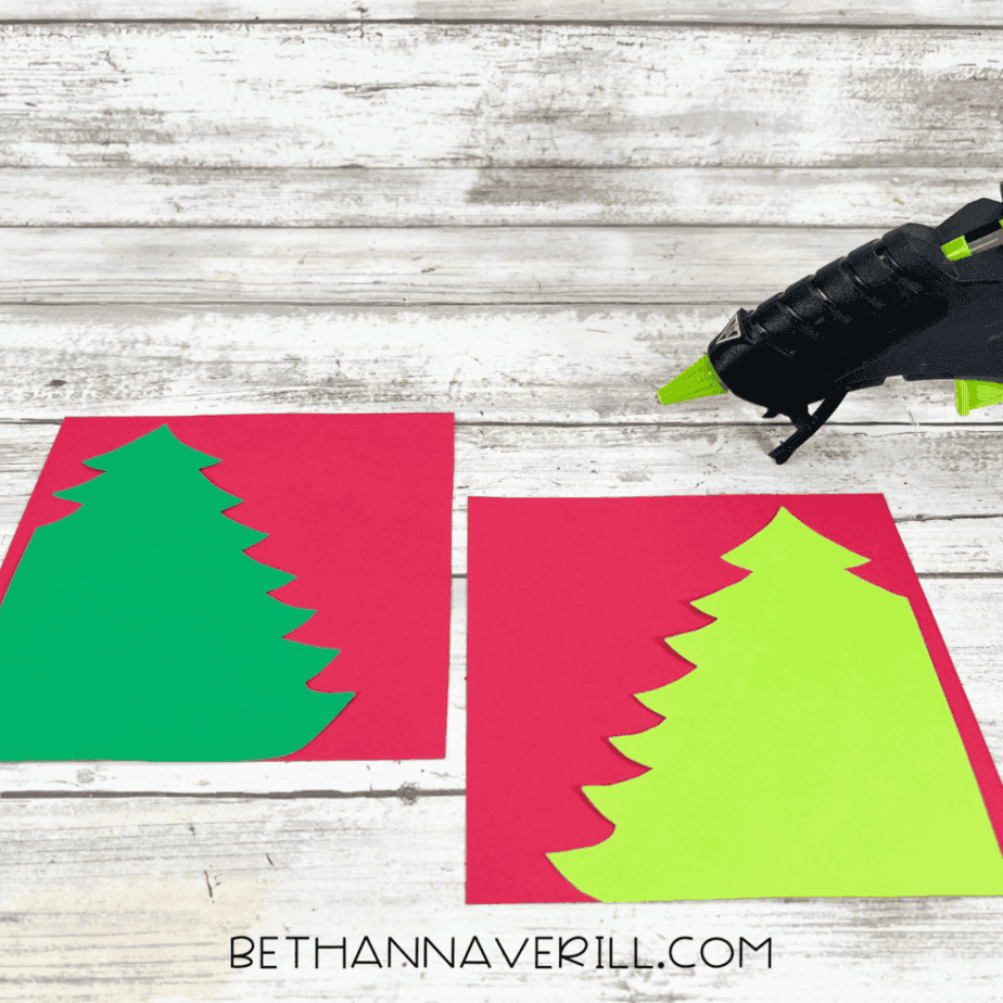 Cut green cardstock Christmas tree shapes placed on red cardstock backgrounds, with a glue gun nearby on a white wood surface.