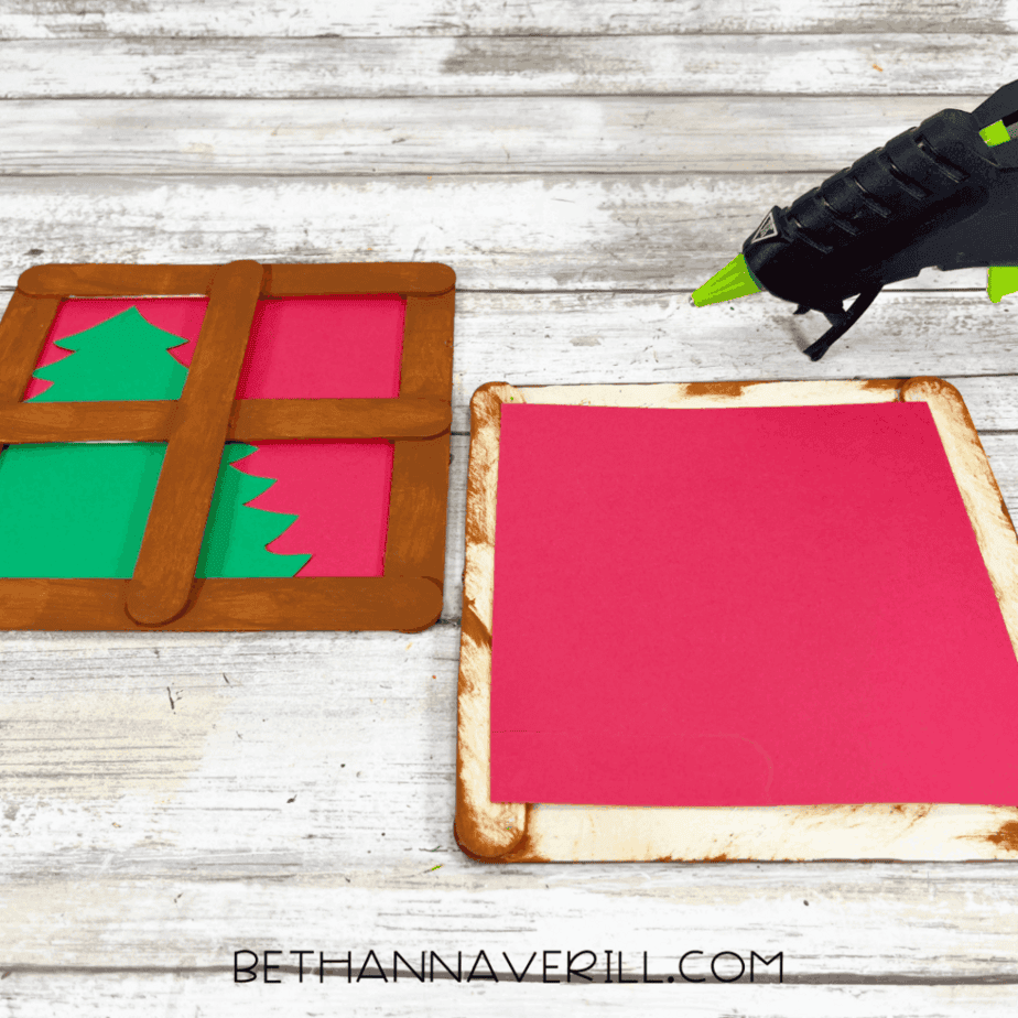 One painted popsicle stick window frame placed over red cardstock with a green Christmas tree underneath, next to another red cardstock square on a white wood background.