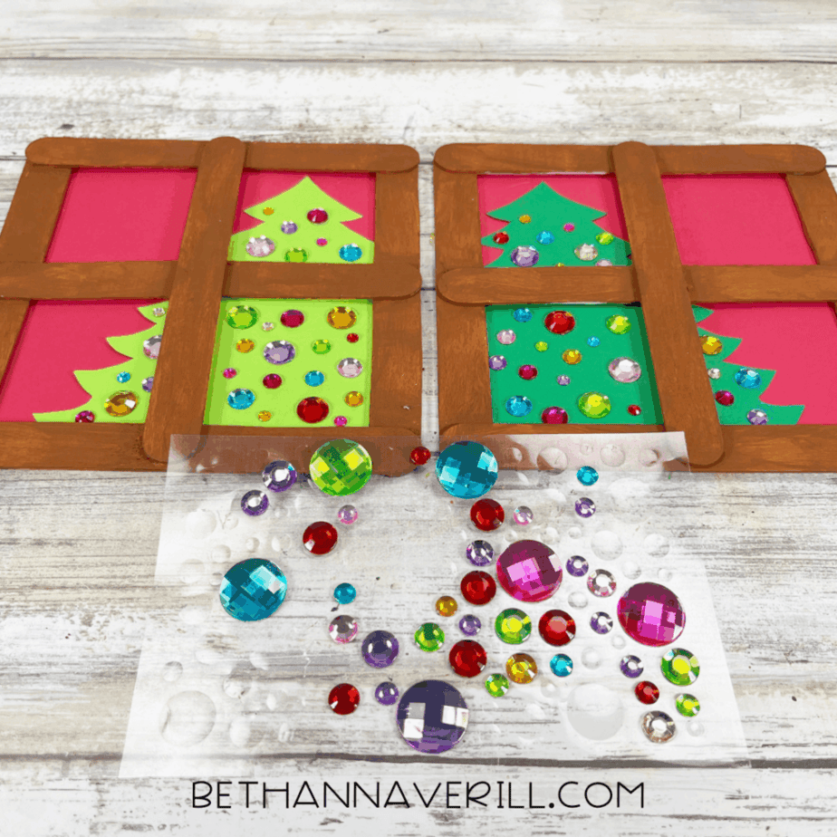 Two Christmas window popsicle stick crafts with green cardstock trees decorated with colorful gem stickers, with extra gems spread out on wax paper in front.
