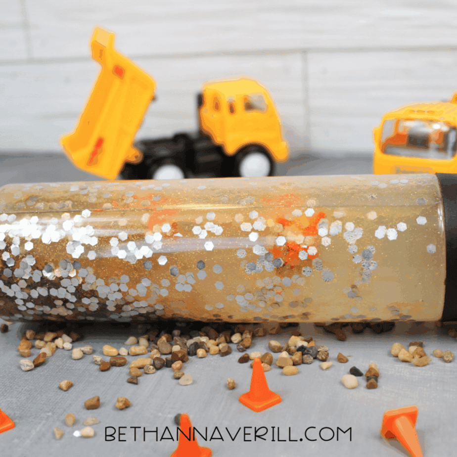 Clear sensory bottle lying horizontally on a gray surface with small pebbles scattered around it and toy construction trucks in the background.