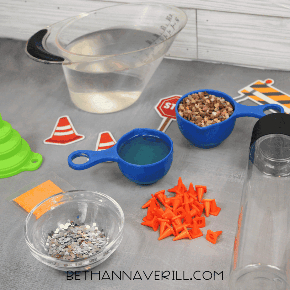 Supplies arranged on a gray surface including a clear plastic bottle, measuring cups with water and small pebbles, orange plastic traffic cones, a small bowl of silver hexagon glitter, and construction-themed cutouts.
