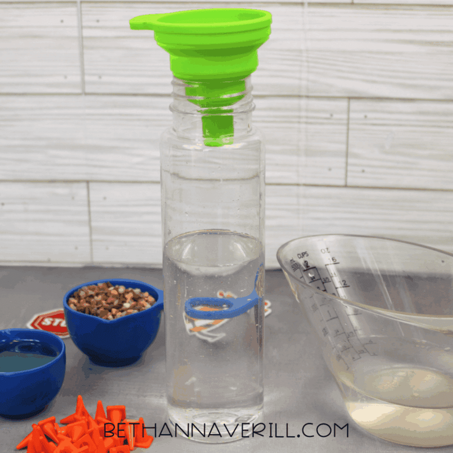 Clear plastic bottle standing upright with a green funnel inserted into the opening, surrounded by measuring cups, small pebbles, and orange plastic traffic cones.