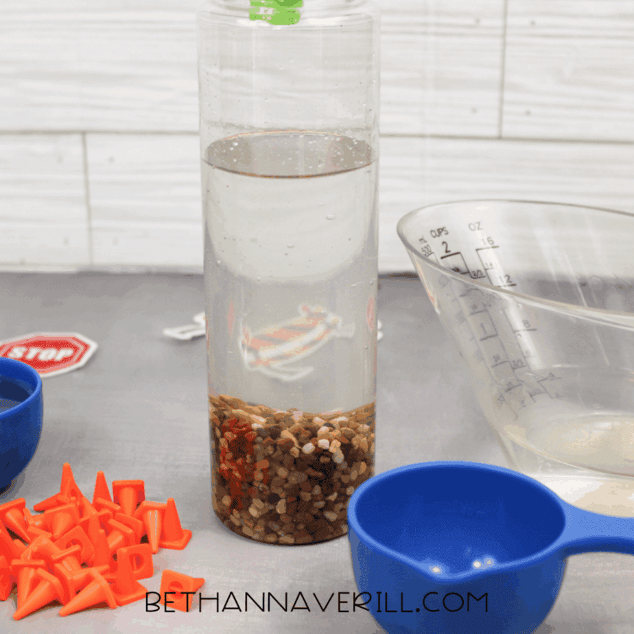 Small pebbles settled at the bottom of a clear plastic bottle partially filled with water, with measuring cups and orange plastic traffic cones nearby.