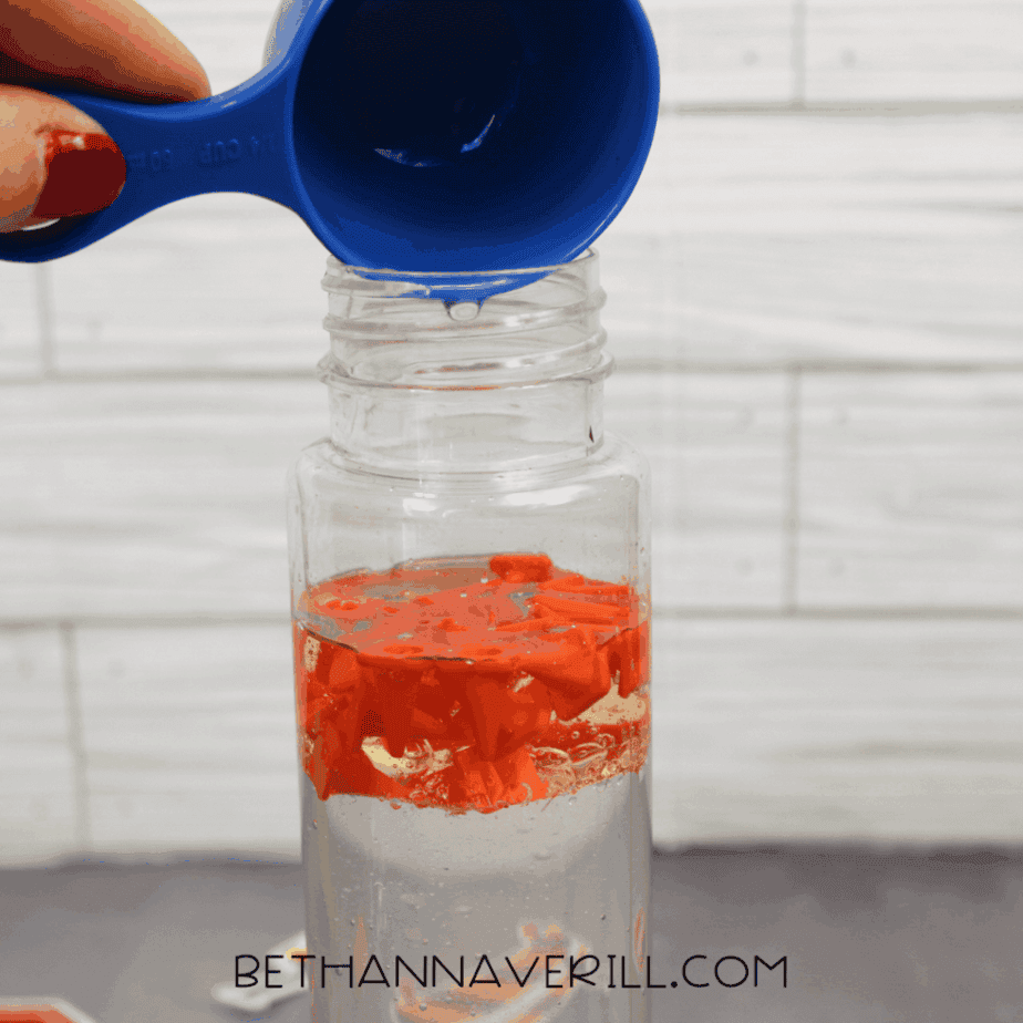 Water being poured from a blue measuring cup into a clear plastic bottle containing orange plastic traffic cones and liquid.