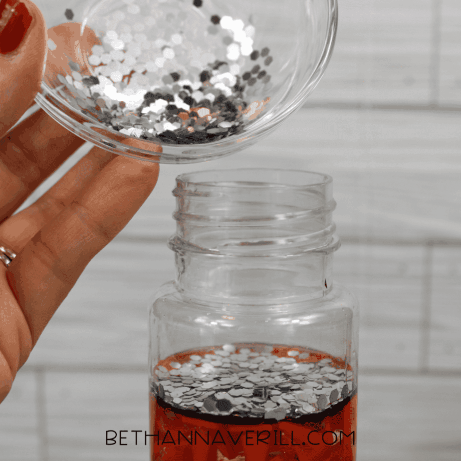 Silver hexagon glitter being poured from a small glass bowl into a clear plastic bottle filled with liquid and orange plastic traffic cones.