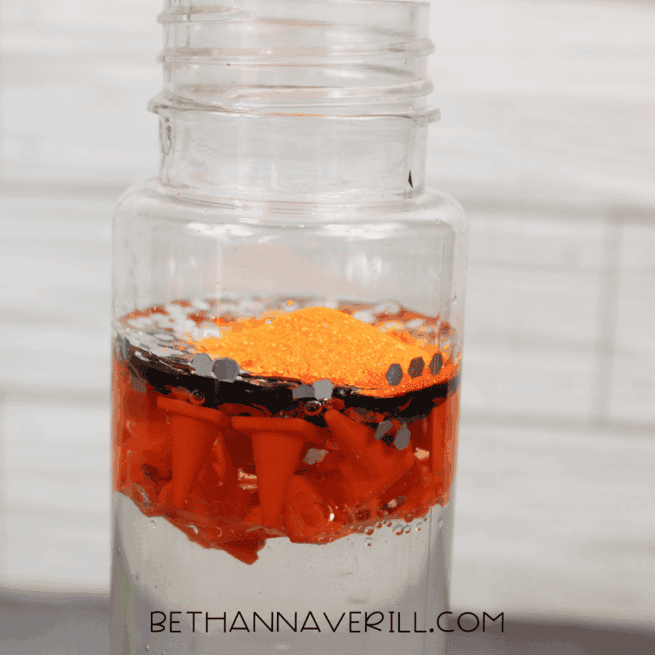 Orange glitter added on top of silver hexagon glitter inside a clear plastic bottle filled with liquid and floating orange plastic traffic cones.