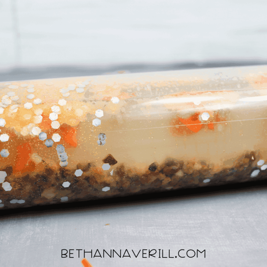 Close-up view of a clear plastic sensory bottle lying on its side, showing glitter, liquid, and small pebbles layered inside.