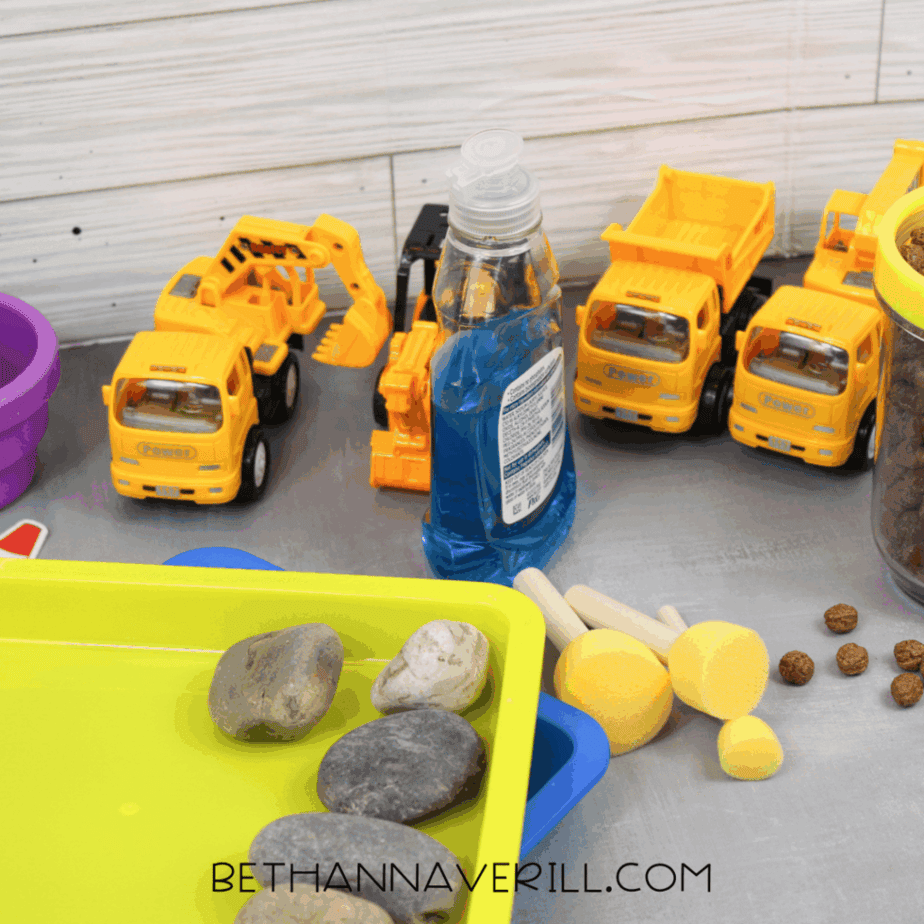 Construction sensory bin setup with toy dump trucks, excavator vehicles, rocks, sand, bubble solution, sponges, and trays arranged on a tabletop.