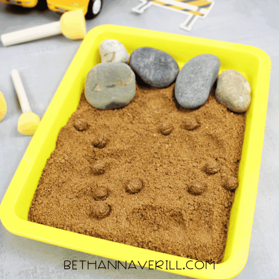 Yellow sensory tray filled with brown sand, smooth rocks, and small dirt clumps for construction-themed sensory play.