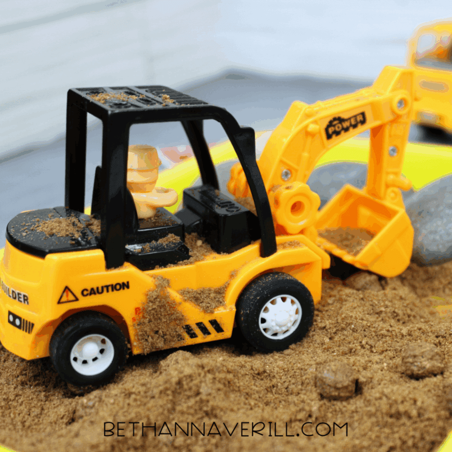 Toy excavator digging into sand inside a yellow sensory tray during construction-themed sensory play.