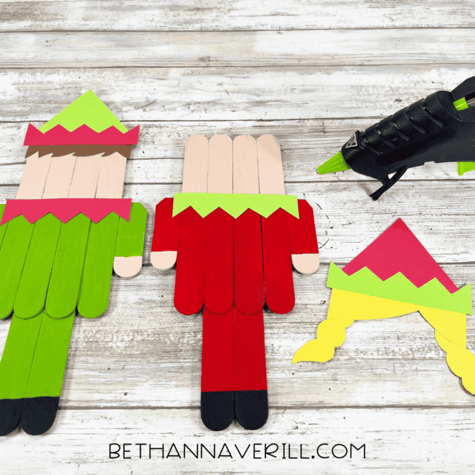 Two popsicle stick elf bodies with cardstock collars and hats placed on top, with additional cardstock pieces and a hot glue gun nearby.