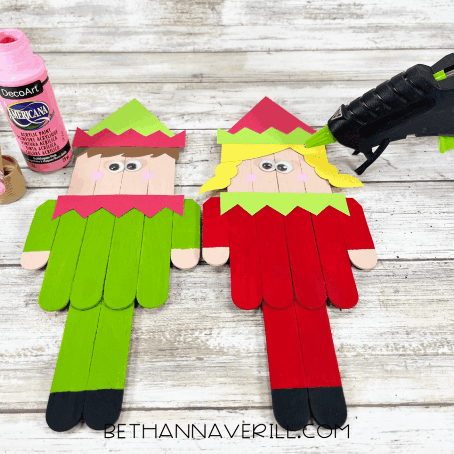Two popsicle stick elves with googly eyes and pink painted cheeks, acrylic paint bottles and a hot glue gun visible on a white wood surface.