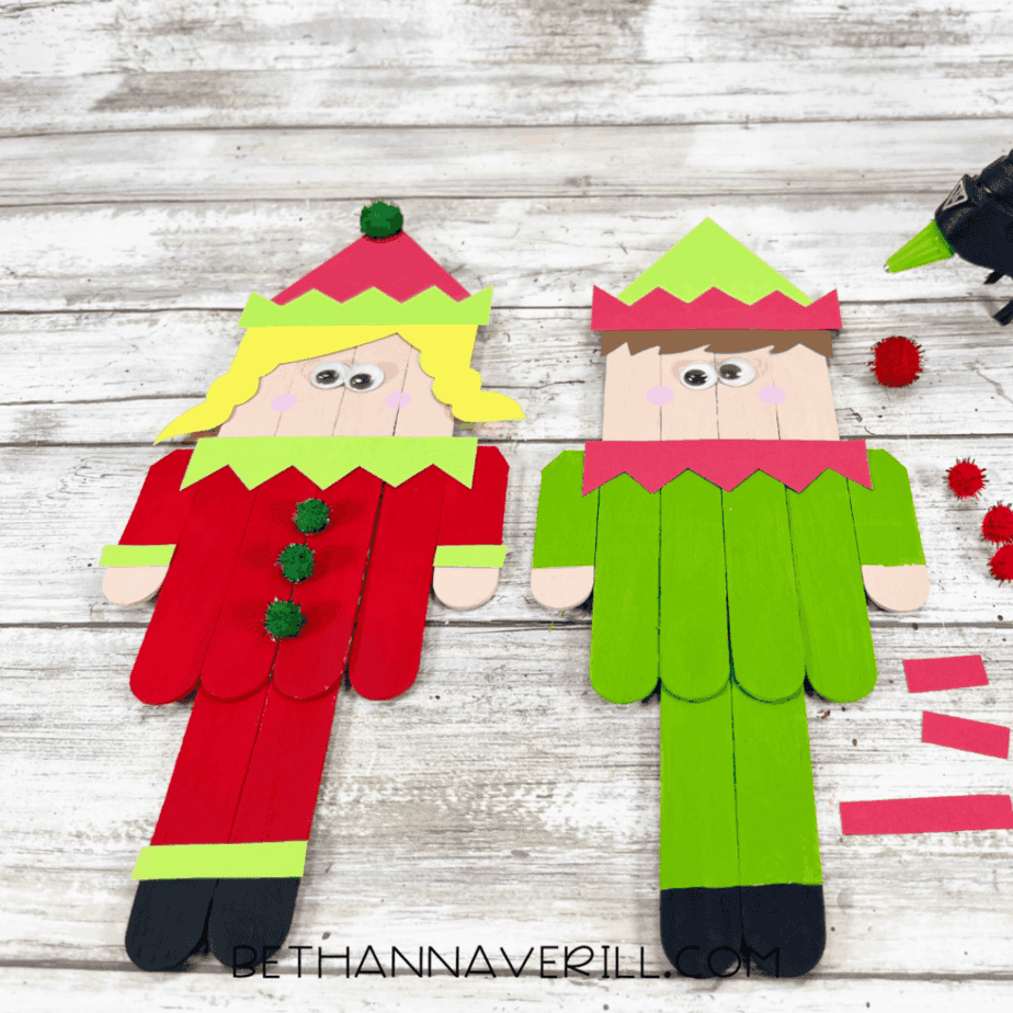 Two popsicle stick elves with cardstock hats and collars, small pom poms placed on the table near the elves and a glue gun to the side.