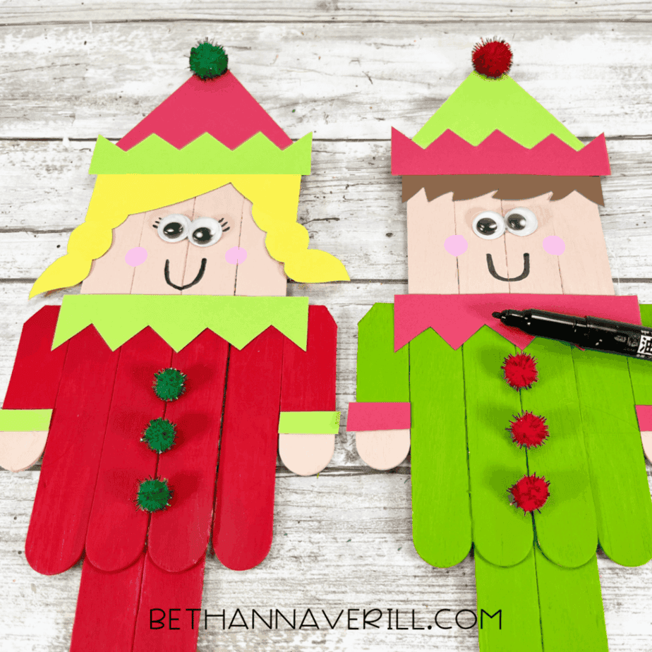 Two popsicle stick elves with drawn smiles, pom poms attached to their bodies, and a black marker resting on the green elf.