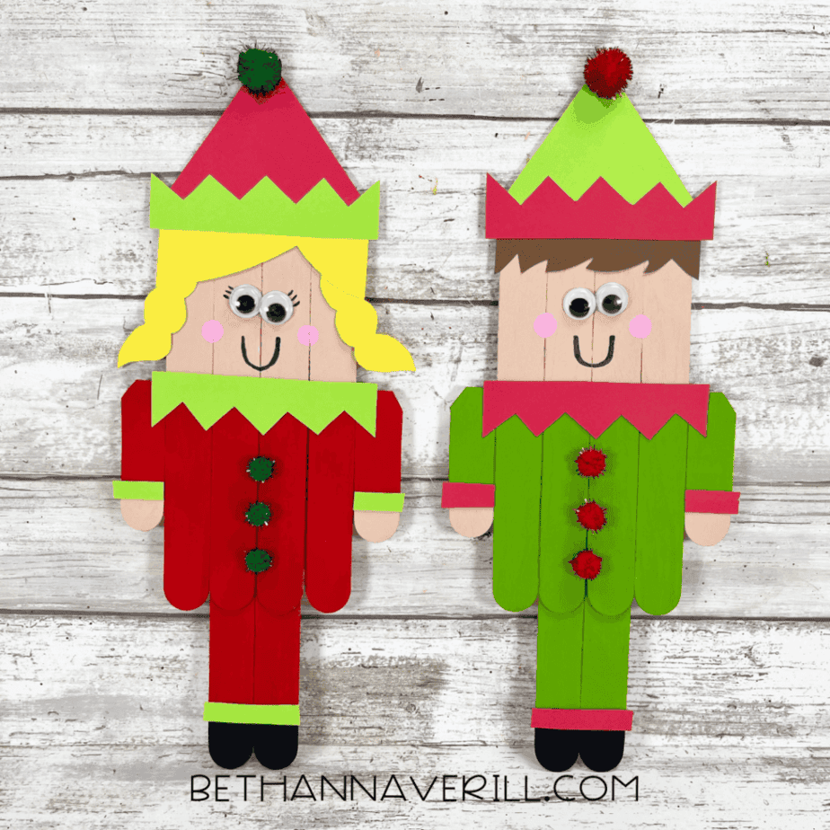 Two completed popsicle stick elf figures displayed side by side on a white wood background.
