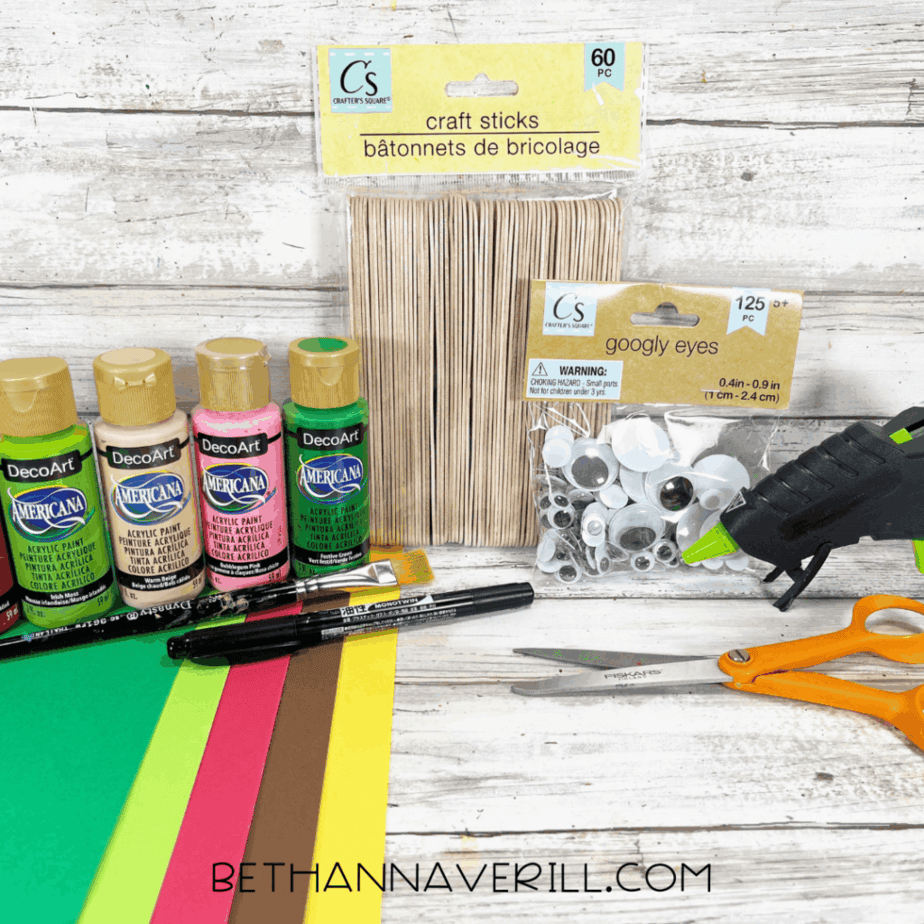 Craft supplies arranged on a white wood surface including craft sticks, googly eyes, acrylic paint bottles, cardstock sheets, a paintbrush, black marker, scissors, and a glue gun.