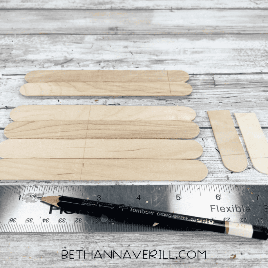 Jumbo craft sticks laid out with pencil markings, a metal ruler, and a pencil on a white wood surface.