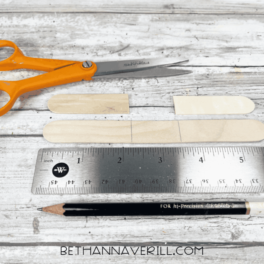 Cut craft stick pieces arranged next to scissors, a ruler, and a pencil on a white wood surface.