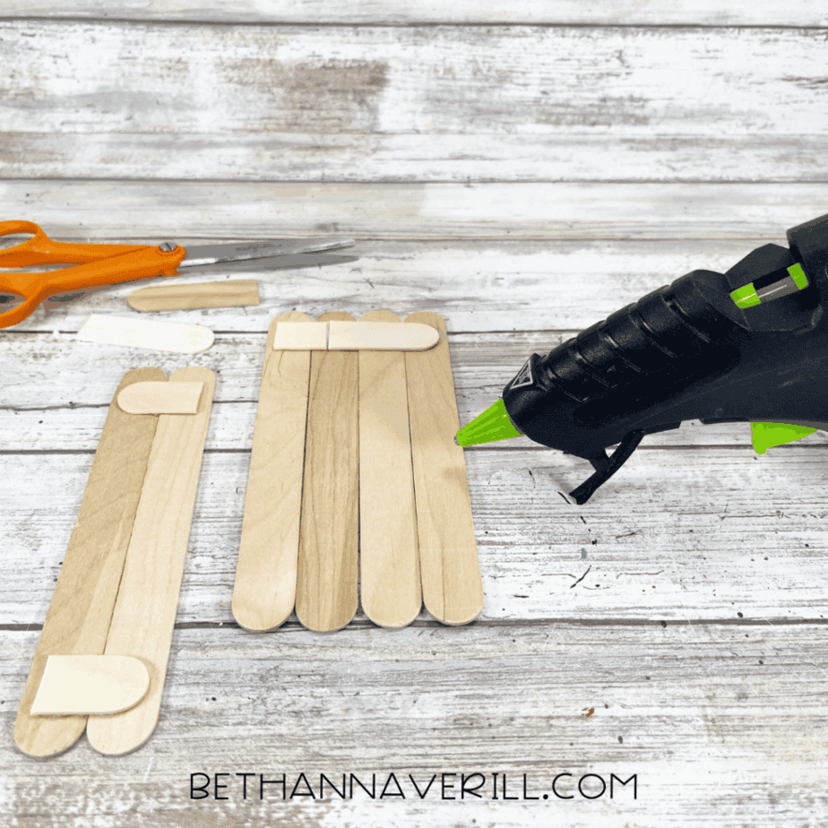 Craft stick pieces arranged into rectangular shapes with smaller stick pieces glued across the top, with a glue gun nearby.