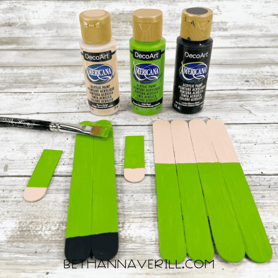 Craft stick pieces partially painted green and skin-tone colors with acrylic paint bottles and a paintbrush visible.