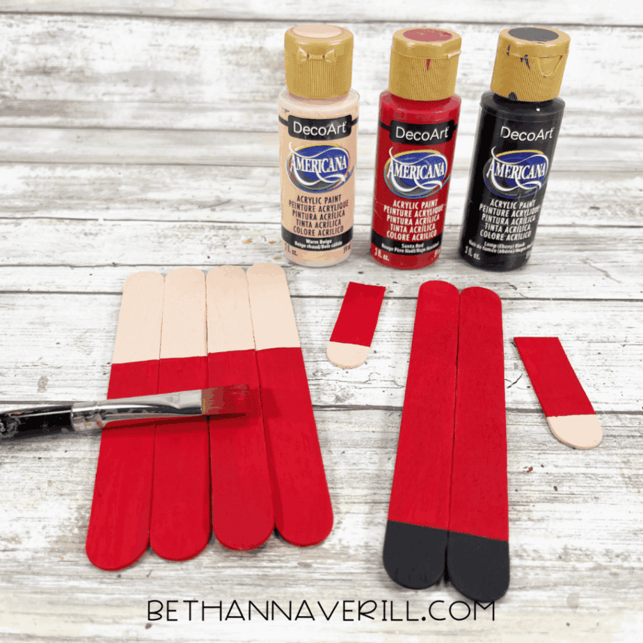 Craft stick pieces partially painted red, black, and skin-tone colors with acrylic paint bottles and a paintbrush visible.