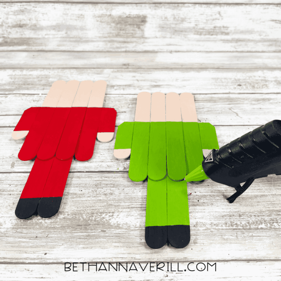 Two popsicle stick elf bodies painted red and green lying on a white wood surface, with a hot glue gun positioned near the green elf.