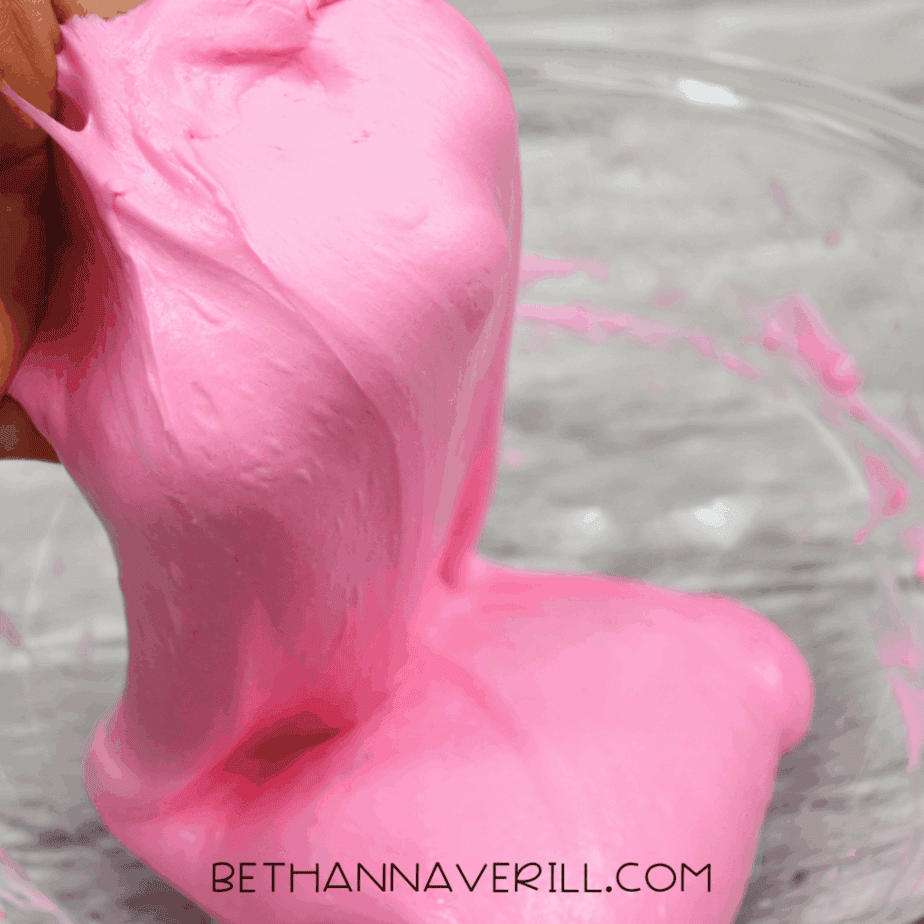 Hand stretching pink fluffy slime above a glass bowl.
