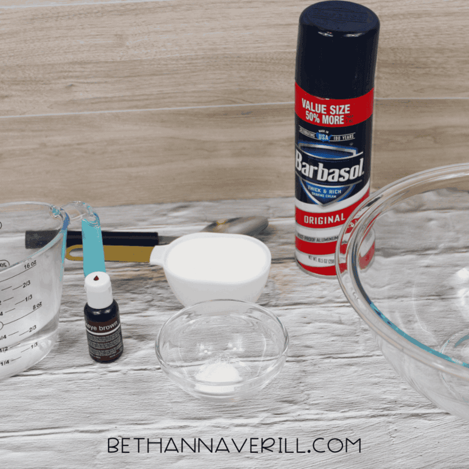 Ingredients and tools arranged on a tabletop including a can of shaving cream, measuring cup with water, small bowl of baking soda, spoon, glass mixing bowl, and bottles of brown and pink food coloring.
