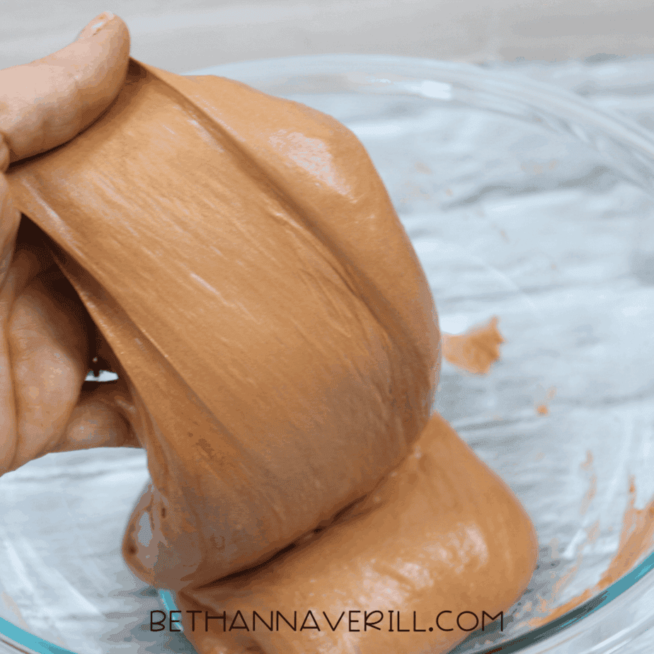 Hand stretching and kneading brown fluffy slime inside a glass bowl.