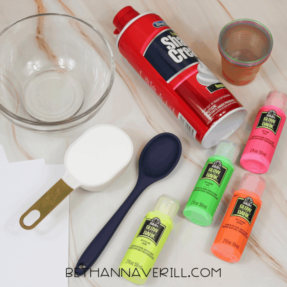 Supplies arranged on a tabletop including a glass bowl, can of shaving cream, plastic cups, measuring cup, spoon, white paper, and bottles of glow in the dark paint in yellow, pink, orange, and green.