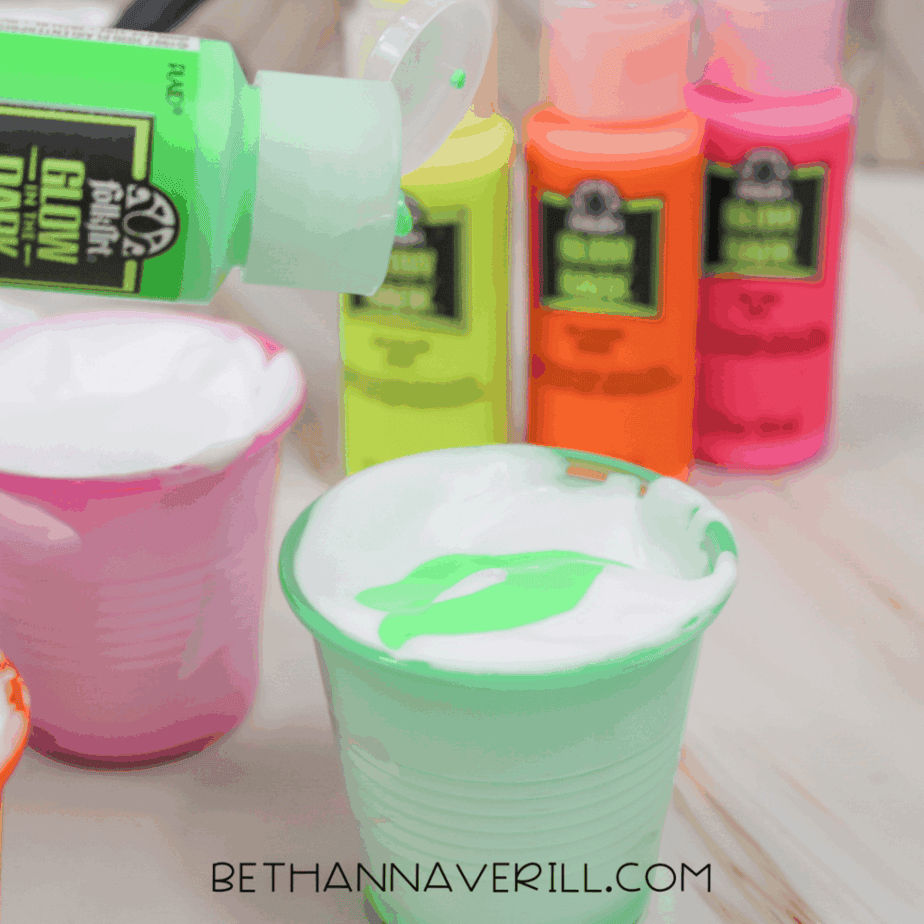Green glow in the dark paint being squeezed into a plastic cup filled with shaving cream, with other glow paint bottles visible in the background.
