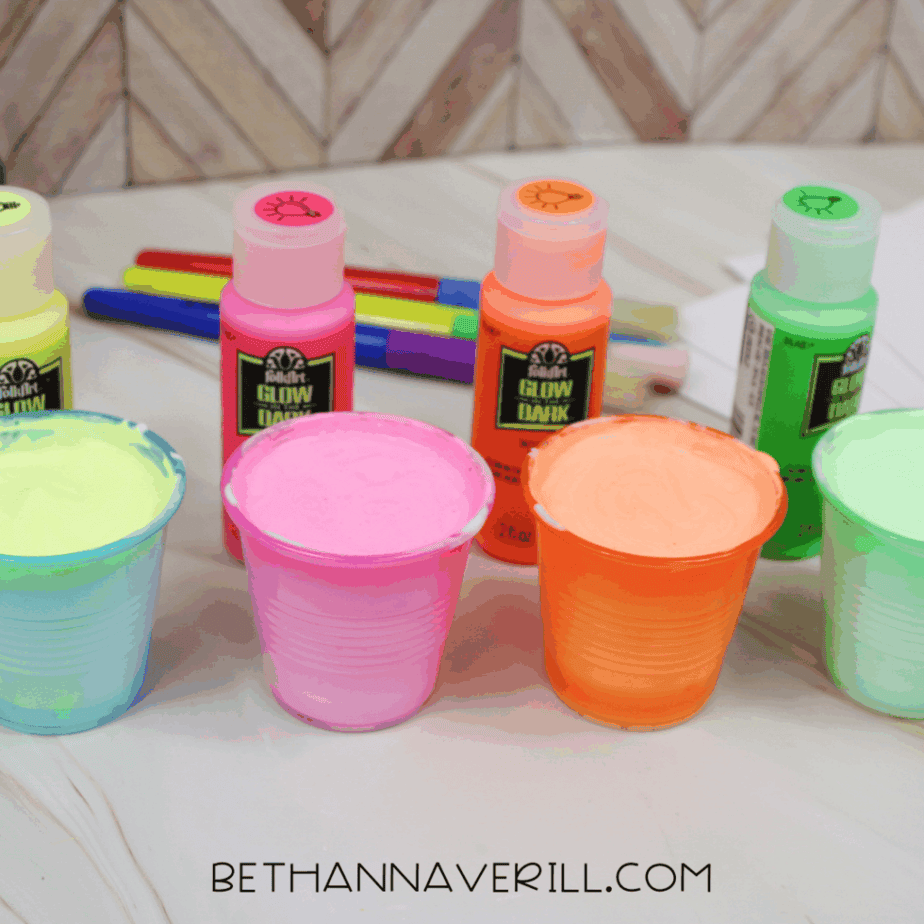 Plastic cups filled with pastel-colored puffy paint mixtures in blue, pink, orange, and green, with glow in the dark paint bottles behind them.