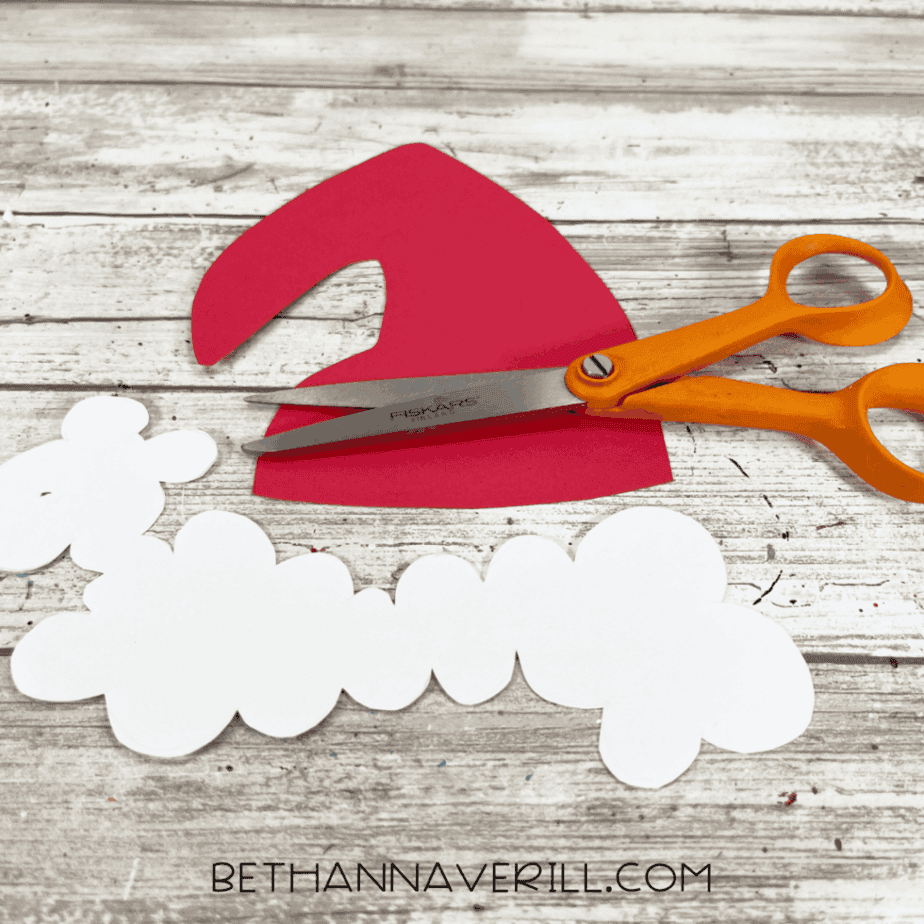 Red cardstock Santa hat shape and white cardstock trim pieces placed on a white wood surface with orange-handled scissors.