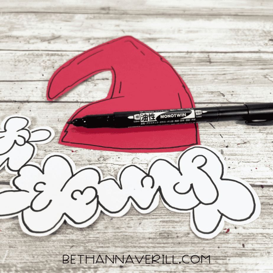 Red cardstock Santa hat shape and white cardstock trim pieces outlined in black marker, with a black marker resting across the hat.