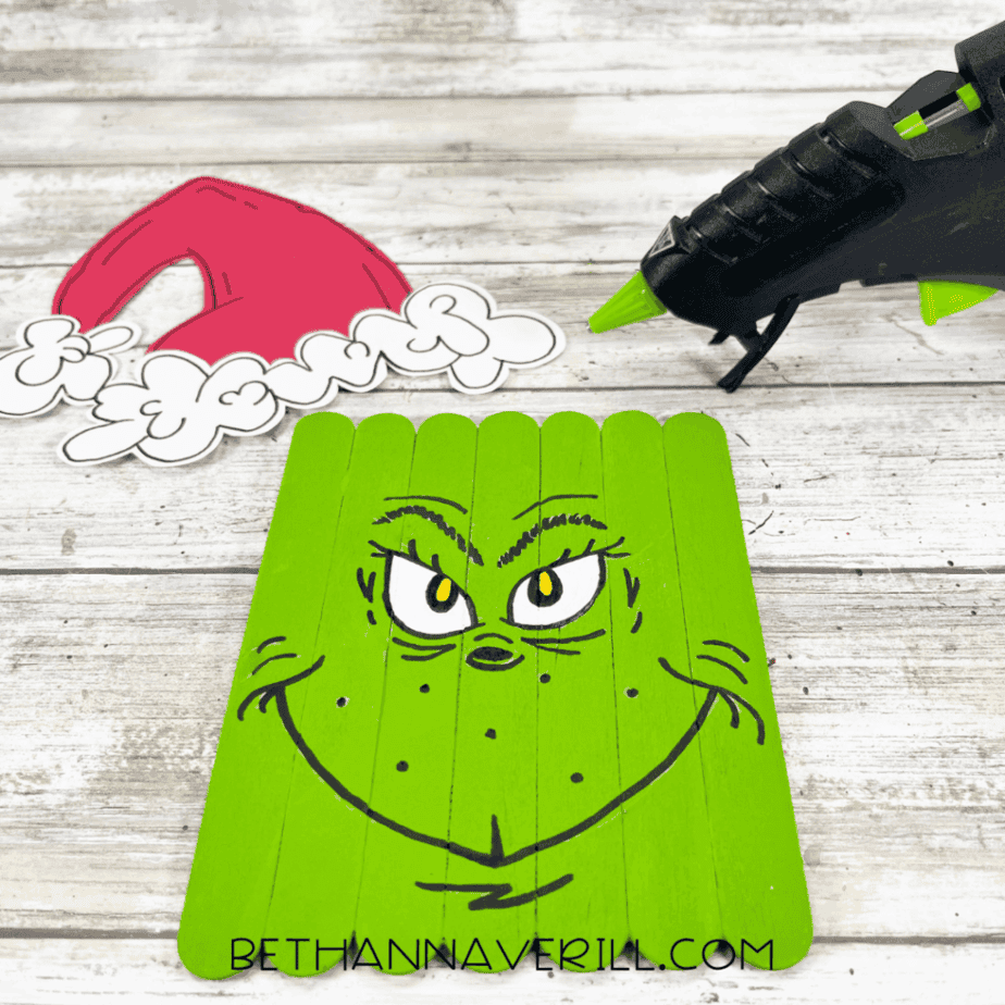 Green-painted craft stick face with black facial details, yellow eyes, a hot glue gun nearby, and paper Santa hat pieces placed above the craft.