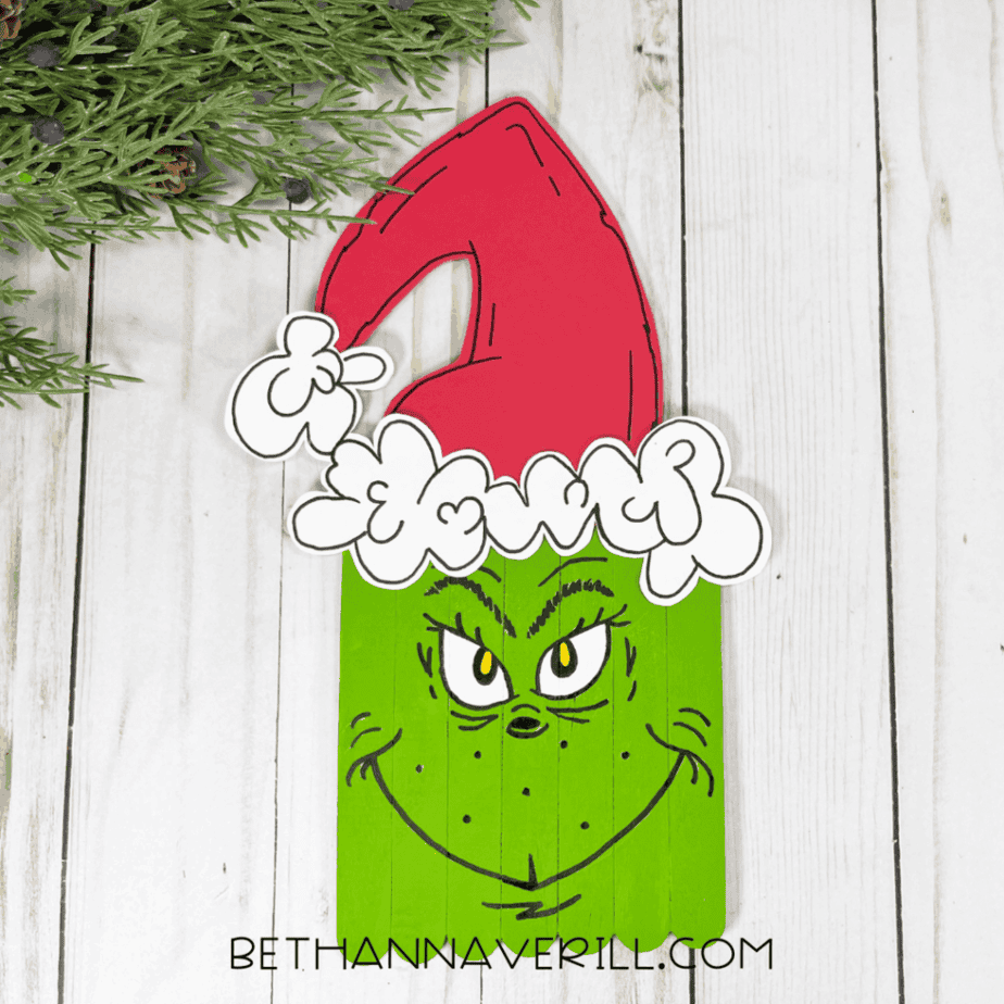 Green-painted craft stick face with a red paper Santa hat and white trim attached, photographed on a white wood surface with greenery in the corner.