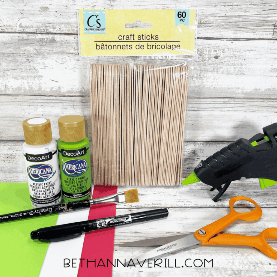 Craft supplies laid out on a white wood surface including a package of craft sticks, green and white acrylic paint bottles, a paintbrush, black marker, scissors, hot glue gun, and sheets of red, white, and green cardstock.