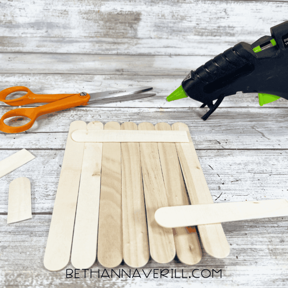 Jumbo craft sticks arranged vertically and glued together with additional craft sticks placed horizontally on the back to hold them together, with scissors and a glue gun nearby.
