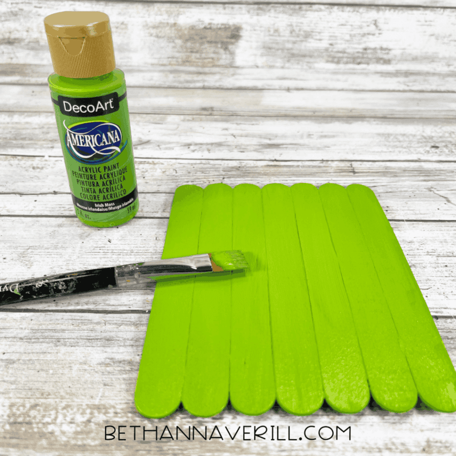 Jumbo craft sticks painted green with a paintbrush resting on top, next to a bottle of green acrylic paint.