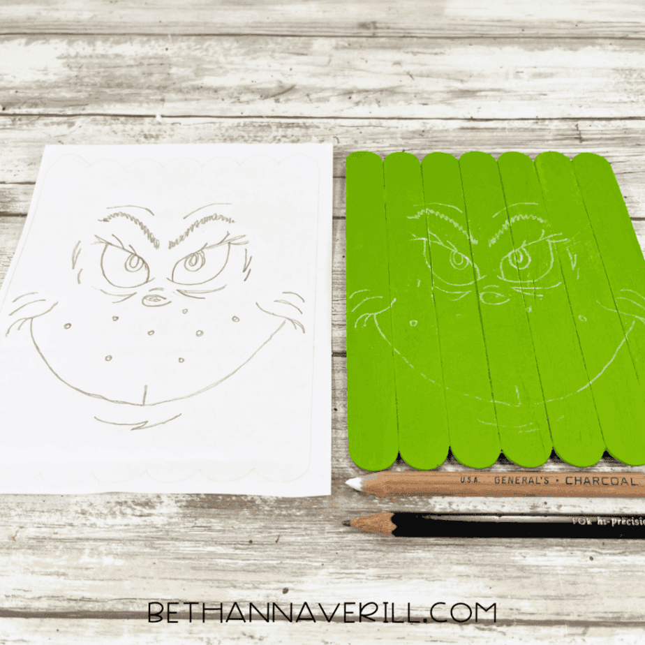 Pencil sketch of a Grinch-style face on white paper next to a green-painted craft stick base with the same face lightly sketched on it using white pencil.