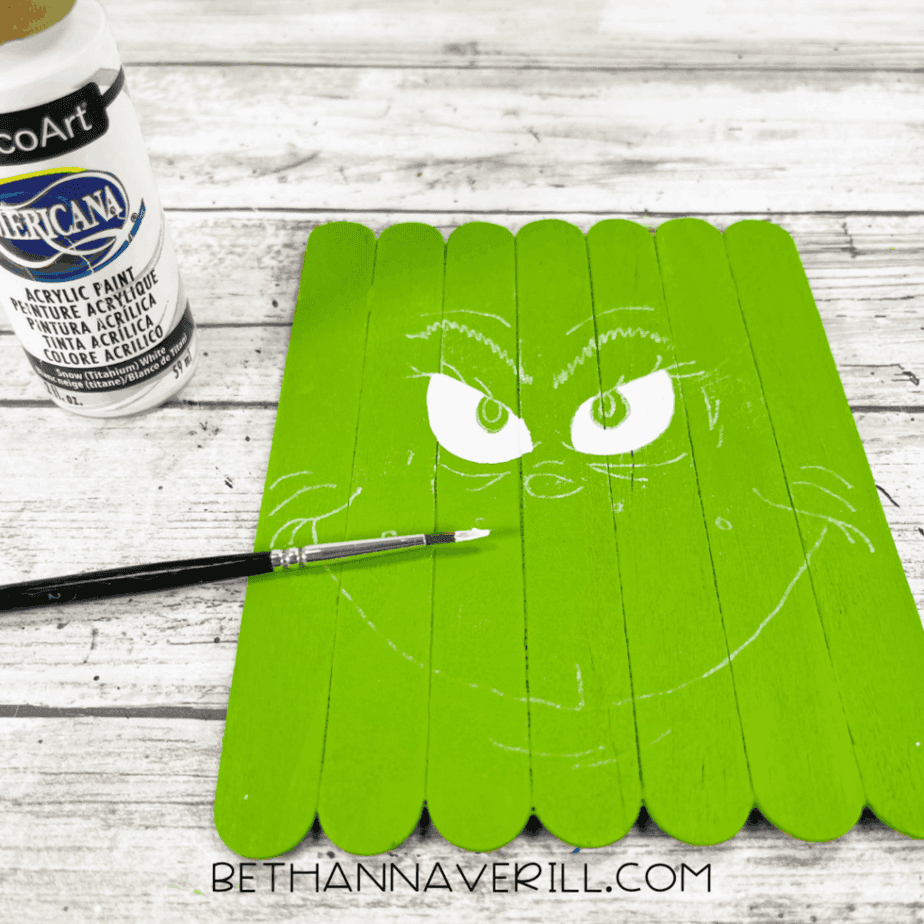 Green-painted craft stick base with white painted eye shapes added, a small paintbrush touching the surface, and a bottle of white acrylic paint nearby.