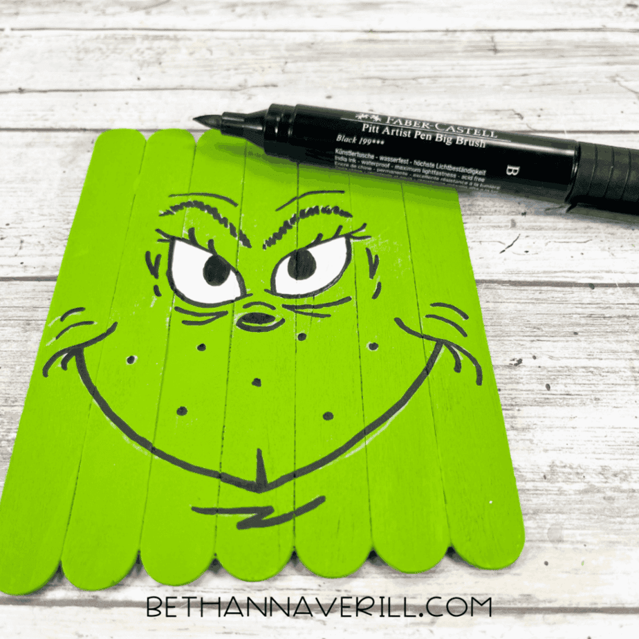 Green-painted craft stick base with black outlines added for eyes, eyebrows, nose, mouth, and facial details, with a black paint pen resting above the craft.