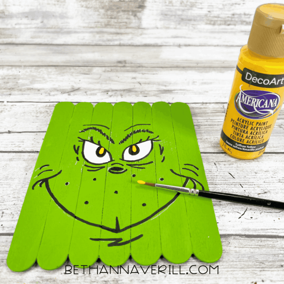 Green-painted craft stick base with yellow details painted inside the eyes, a small paintbrush touching the surface, and a bottle of yellow acrylic paint nearby.