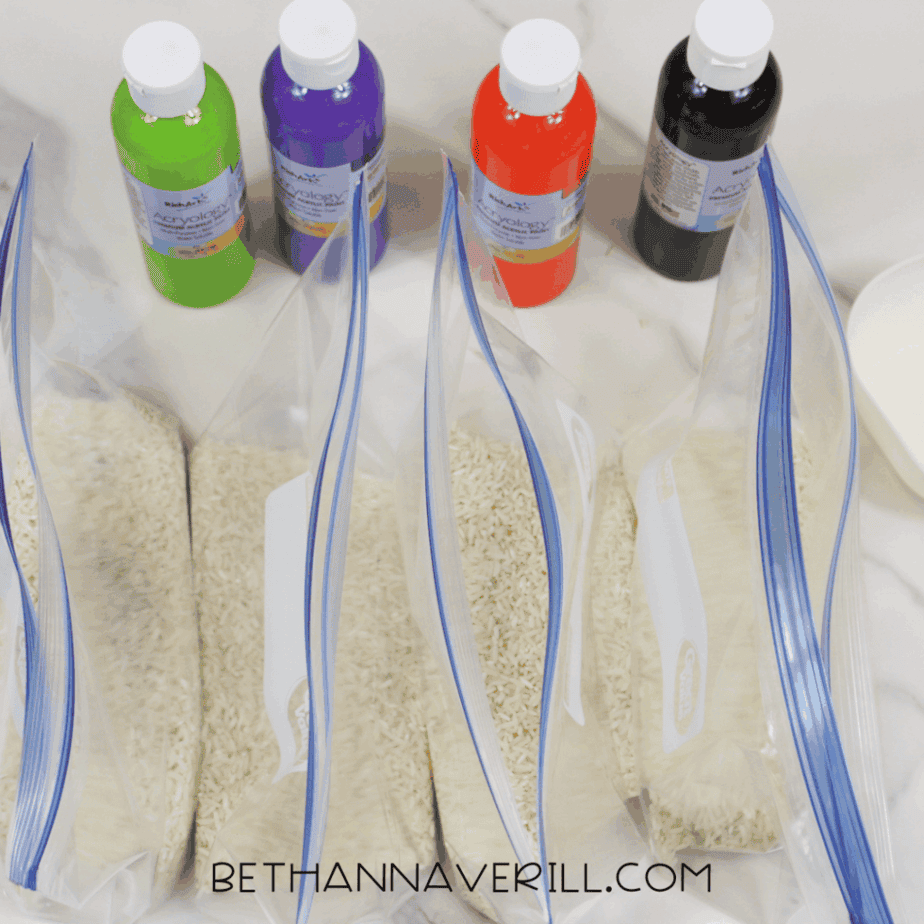 Four resealable plastic bags filled with white rice lined up beneath bottles of green, purple, orange, and black acrylic paint before coloring.