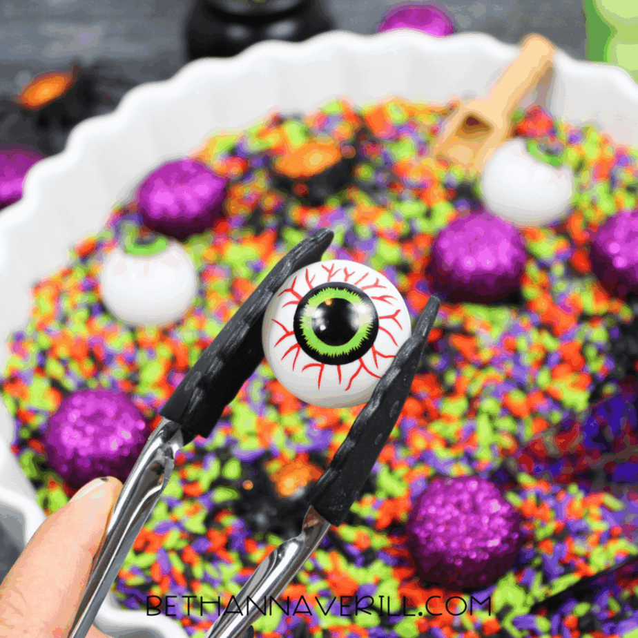 Tongs holding a plastic eyeball above a Halloween rice sensory bin filled with colorful rice and themed manipulatives.