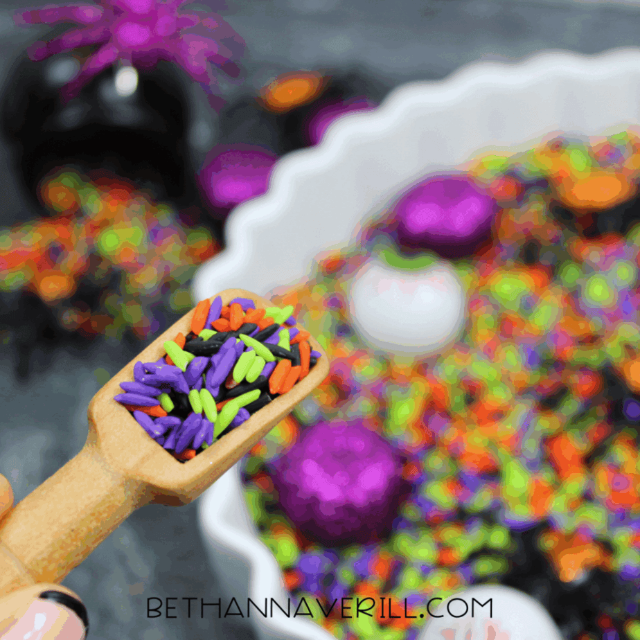 Wooden scoop filled with green, purple, orange, and black rice lifted above a Halloween sensory bin.
