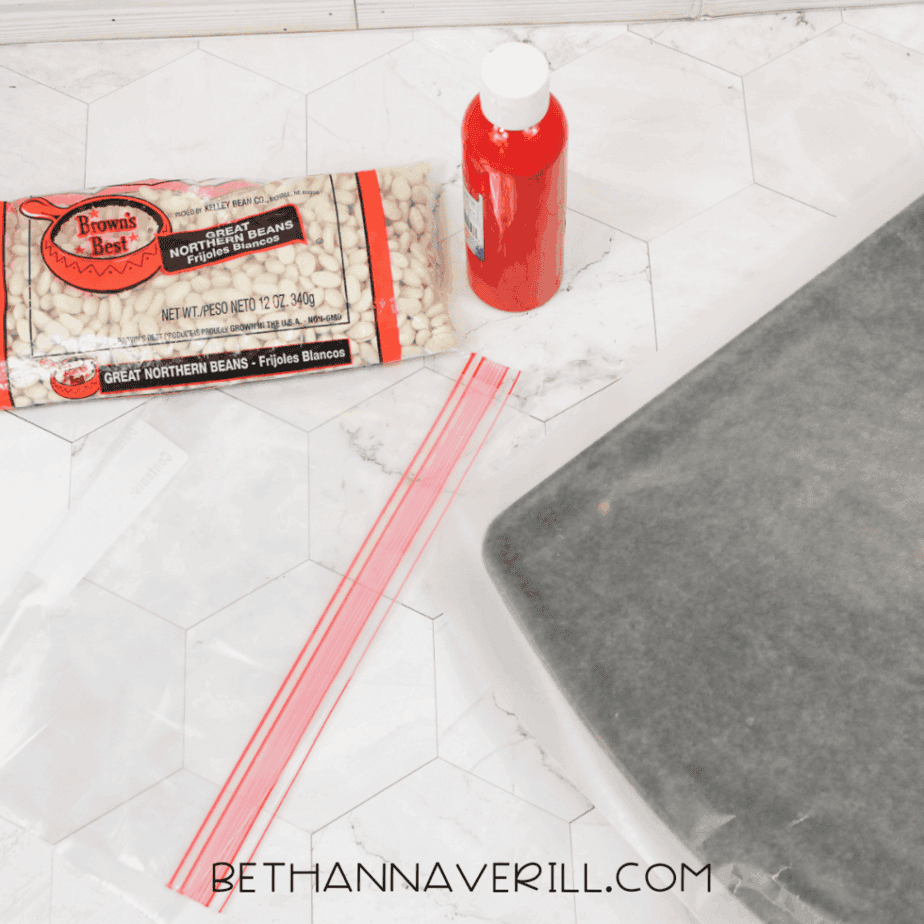 Supplies for a ladybug sensory bin including white beans, red acrylic paint, zip-top bags, wax paper, and a baking sheet on a countertop.