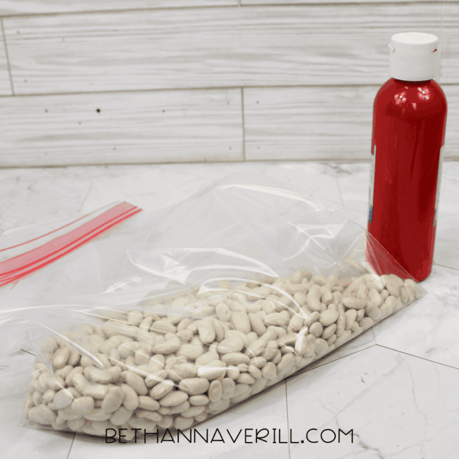 Dried northern white beans inside a resealable plastic bag next to a bottle of red acrylic paint before coloring for a sensory bottle.