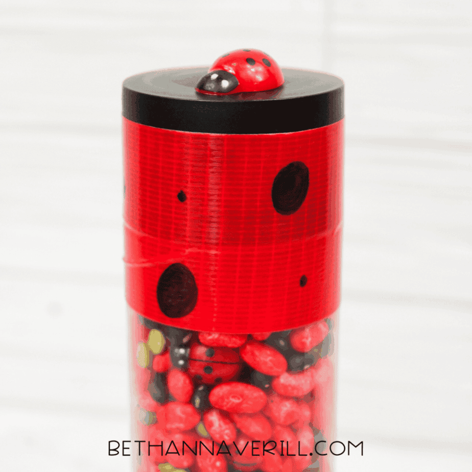 Plastic ladybug toys scattered next to a clear sensory bottle filled with red, black, and green dried beans.